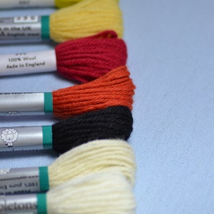 May include: Close-up of a selection of colorful embroidery floss spools. The spools are in various colors, including black, yellow, red, orange, and cream. The floss is labeled as 100% wool and made in England.