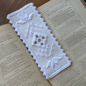 May include: White embroidered bookmark with a scalloped edge and floral designs. The bookmark is lying on top of an open book.