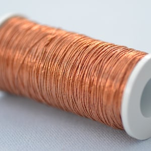 Puede incluir: Una bobina de hilo metálico de color cobre enrollada alrededor de una bobina de plástico blanco.