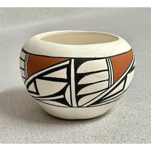 Può includere: Una piccola ciotola in ceramica di forma rotonda color bianco sporco, caratterizzata da un motivo geometrico dipinto a mano in nero opaco e rosso terracotta. Il design presenta linee angolari, triangoli e segmenti curvi sulla superficie.