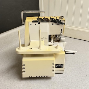 Puede incluir: Una remalladora Singer de color crema y blanco con asa de transporte metálica, tres portacarretes verticales y panel de control frontal. La máquina muestra el número de modelo 14U 52A y el texto Made in Japan en su carcasa.