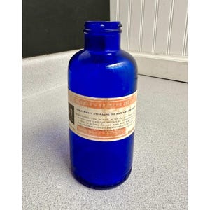 May include: A vintage cobalt blue glass bottle with a narrow neck and a wide base. A rectangular paper label is wrapped around the centre, featuring the text Blumer's Shampoo Jelly, directions for use, and the location Chicago, Illinois.
