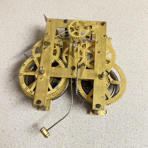 May include: A brass clock movement mechanism featuring an intricate assembly of interlocking gears, wheels, and plates. The mechanical device includes a thin metal wire with a small cylindrical weight attached, showcasing exposed internal components.