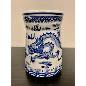 May include: A cylindrical ceramic vessel featuring a white base with blue hand-painted details. The design displays a large dragon motif surrounded by stylized clouds and waves, with a decorative geometric border pattern around the top rim.