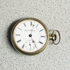 May include: A gold-tone pocket watch with a white dial, black Roman numeral hour markers, and a small sub-dial for seconds. The face features the text Elgin Nat Watch Co in black script. It includes a winding crown and a circular top loop.