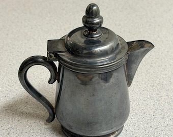 Puede incluir: Una jarra de metal plateado pulido o tetera pequeña con asa curva, tapa con bisagra y pomo decorativo, y un pico vertedor corto. El recipiente tiene una superficie lisa y reflectante con un diseño de base redondeada.