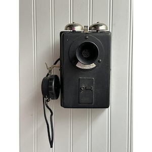 May include: A vintage black wall-mounted telephone featuring a rectangular housing, a circular mouthpiece, and two silver-toned metal bells at the top. A black circular earpiece is attached to the side by a black cord, hanging from a metal hook.