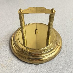 May include: A circular gold-toned metal base featuring a smooth, reflective surface with a central small cylindrical post. Two vertical textured pillars support a horizontal rectangular top plate with two small holes at the center.