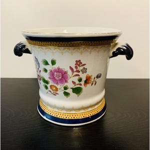 Puede incluir: Un macetero de cerámica blanca con un estampado floral de flores rosas, naranjas y azules. El recipiente tiene un borde y una base de color azul oscuro con adornos dorados y dos asas de color azul oscuro con forma de voluta.