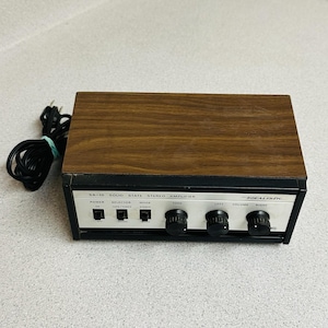 May include: A Realistic SA-10 solid state stereo amplifier with a wood grain top and black metal base. The front silver control panel features a power switch, selector and mode switches, and three black rotary knobs for tone, left volume, and right volume. A black power cord is attached.