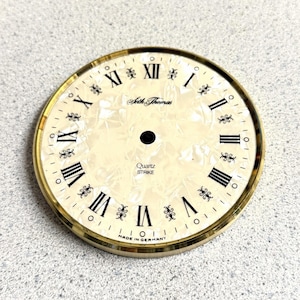 May include: A round clock dial with a cream-colored, pearlescent finish and a gold-toned metal rim. It features black Roman numerals, decorative flourishes between the numbers, and the text Seth Thomas, Quartz STRIKE, and MADE IN GERMANY.