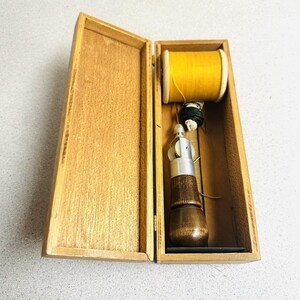 May include: A rectangular wooden box containing a sewing awl with a dark brown wooden handle and a silver metal tip. Inside the box, there is also a spool of bright yellow thread and a small bundle of black thread, all arranged neatly within.