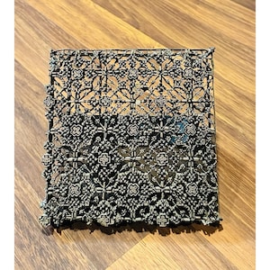 May include: A square, dark metal block with an intricate, repeating floral and geometric pattern. The design features cross-like shapes and delicate lines, creating a detailed, openwork appearance. The block is likely used for printing or decorative purposes.