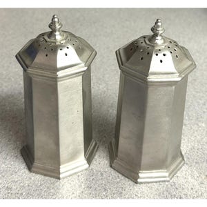 May include: Two matching silver-colored salt and pepper shakers. Each shaker has an octagonal body and a decorative, pointed finial on top. The tops have small holes for dispensing. The shakers are made of a shiny, metallic material.