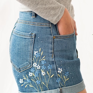 May include: Light blue denim shorts with a rolled hem. The shorts feature an embroidered floral design in shades of blue and white, with yellow stems. White speckles are scattered across the denim.