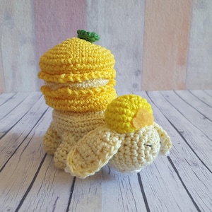 Puede incluir: Un conejito de crochet amarillo y crema con un macaron amarillo en la espalda. El conejito está acostado sobre una superficie de madera blanca.