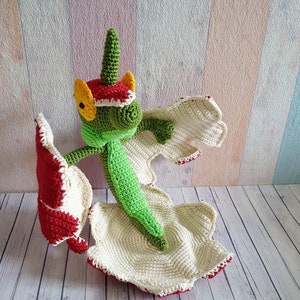 Puede incluir: Un juguete de dragón de crochet verde y rojo con una concha blanca y crema. El dragón tiene un ojo amarillo y una cola larga verde.