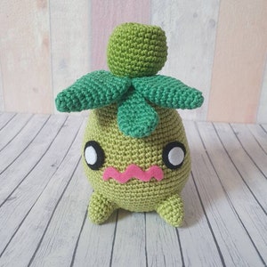 Könnte beinhalten: Grünes Häkel-Plüschtier mit einem runden Kopf, großen Augen und einem rosa Mund. Das Spielzeug hat einen grünen, blattartigen Kopfschmuck.
