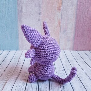 May include: A crocheted purple cat sitting on a white wooden surface. The cat has a long tail and is facing away from the camera.