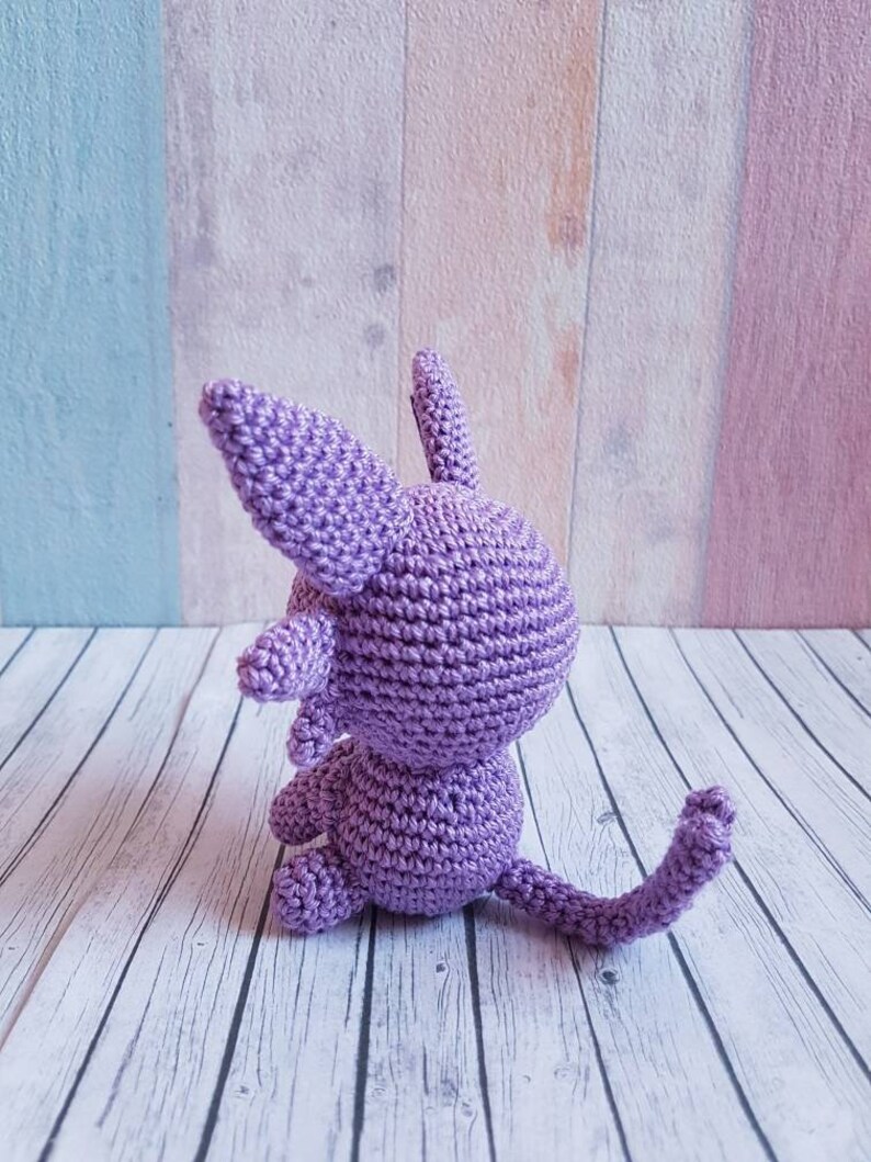 May include: A crocheted purple cat sitting on a white wooden surface. The cat has a long tail and is facing away from the camera.