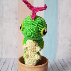 Puede incluir: Un juguete de oruga de crochet verde y amarillo con un tallo rosa en la parte superior, sentado en una maceta de terracota.