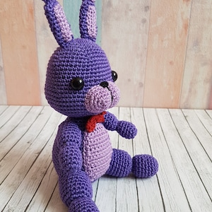 Puede incluir: Juguete de peluche de conejo de crochet morado con ojos negros y una pajarita roja. El juguete está sentado sobre una superficie de madera blanca.