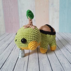May include: A crocheted green turtle with a small green tree growing on its back. The turtle has yellow accents on its legs and head. The turtle is sitting on a white wooden surface.