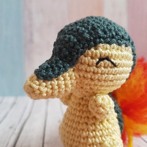Puede incluir: Un juguete de peluche tejido a crochet que se asemeja a un personaje de Pokemon. El juguete es de color crema con una cabeza verde oscuro y una cola esponjosa naranja y amarilla.