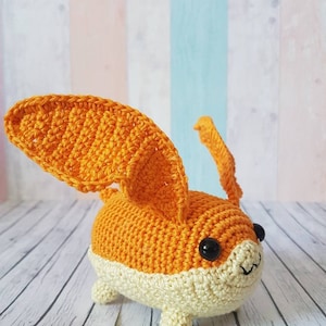May include: A crocheted orange and cream colored stuffed animal with large ears and black eyes. The animal is sitting on a white wooden surface.