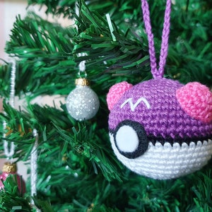 May include: A purple and white crocheted Poké Ball ornament with pink ears, hanging on a green artificial Christmas tree.