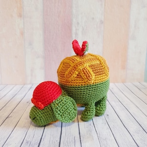 Puede incluir: Dos juguetes de tortuga de crochet, uno más grande que el otro. La tortuga más grande es amarilla, naranja y verde, con un corazón rojo en la espalda. La tortuga más pequeña es verde con un sombrero rojo.