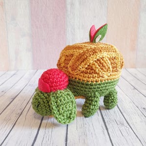 Pode incluir: Dois peluches de tartaruga de crochê, um maior que o outro. A tartaruga maior é verde e amarela com uma folha vermelha no topo. A tartaruga menor é verde com uma flor vermelha no topo.