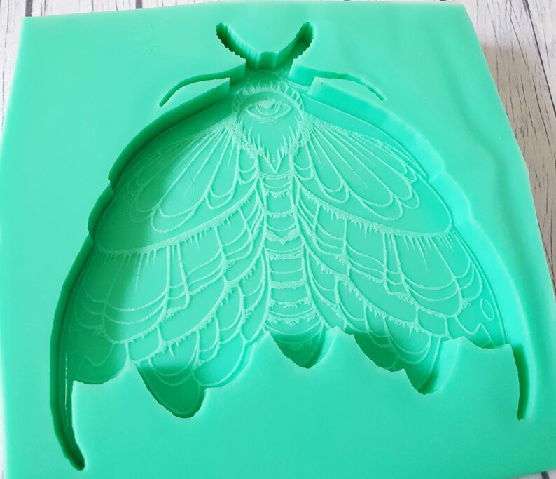Puede incluir: Un molde de silicona verde con forma de polilla con alas detalladas y un gran ojo en el centro.