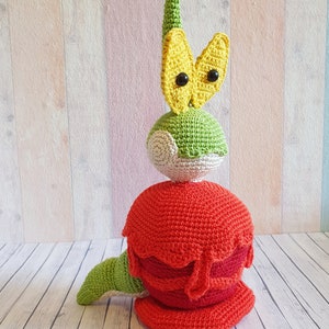 Puede incluir: Un juguete de peluche tejido a crochet con forma de criatura verde y roja con una cabeza amarilla y ojos grandes. El juguete está de pie sobre una superficie de madera.