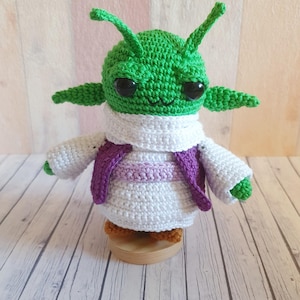 May include: A handmade green crochet alien doll with large black eyes, antennae, and small arms. The alien is wearing a white outfit with a purple vest. It is displayed on a wooden stand.