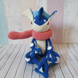 May include: A blue and white crocheted plush toy with a red scarf. The toy is sitting on a wooden surface.