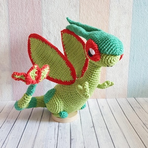 Puede incluir: Un juguete de dragón de crochet verde y rojo con una cola larga y alas grandes. El dragón tiene ojos rojos y un cuerpo verde con detalles rojos.