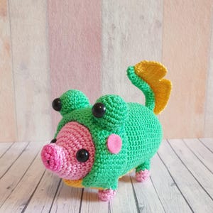 Könnte beinhalten: Ein grünes, gehäkeltes Stofftier mit einer rosa Schnauze und einem gelben Schwanz. Das Tier hat große schwarze Augen und einen rosa Kreis auf dem Kopf.
