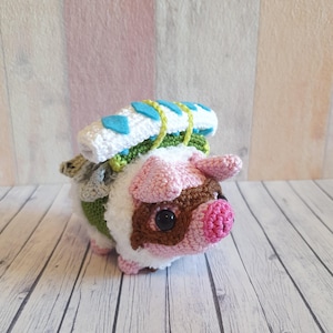 Op de afbeelding: Een gehaakt roze varken dat een groen-witte rugzak draagt met blauwe accenten. Het varken heeft bruine ogen en een roze snuit.