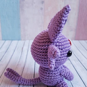 May include: A crocheted purple plush toy with a long tail and large ears. The toy has black eyes and a red nose.