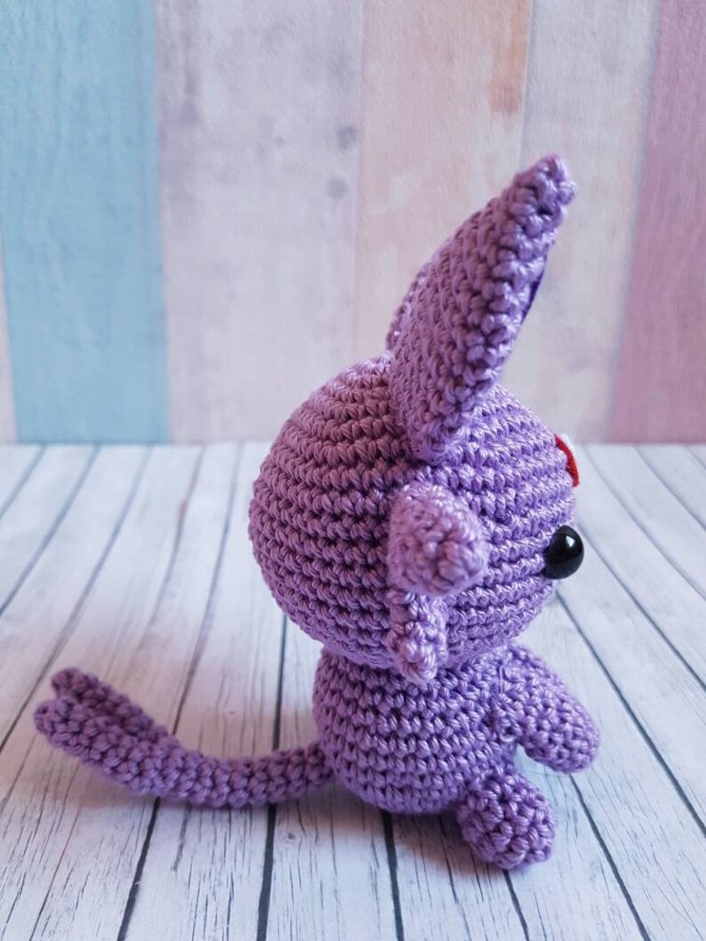 May include: A crocheted purple plush toy with a long tail and large ears. The toy has black eyes and a red nose.