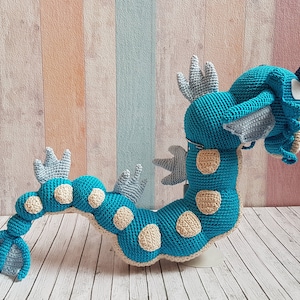 May include: A crocheted blue and beige dragon with a long tail and a spiky head. The dragon is standing on a white surface.