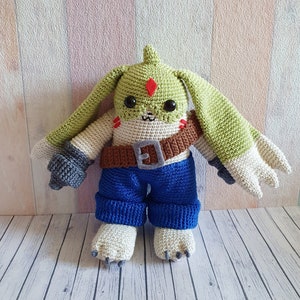 Peut inclure: Un jouet en peluche crocheté avec des oreilles vertes, un corps blanc, un pantalon bleu et une ceinture marron. Le jouet a un diamant rouge sur le front et des yeux noirs.