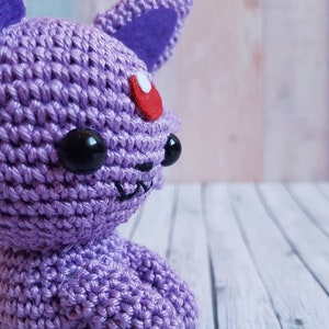 May include: A crocheted purple Eevee plush toy with black eyes and a red gem on its forehead. The toy is sitting on a white wooden surface.
