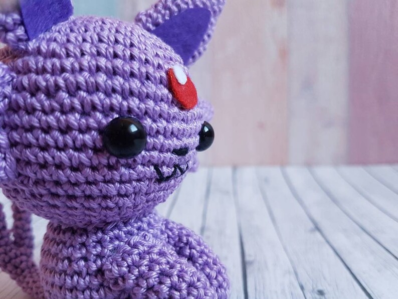 May include: A crocheted purple Eevee plush toy with black eyes and a red gem on its forehead. The toy is sitting on a white wooden surface.