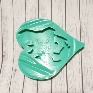 May include: A green heart-shaped silicone mold with a design of a cartoon character with a white outline. The character has a blue and white color scheme and is in a fighting pose.