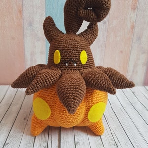 May include: A crocheted plush toy in the shape of a brown and orange creature with a curled tail and yellow eyes.