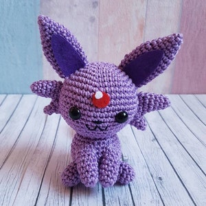 May include: Purple crocheted plush toy resembling a Pokemon character with large ears, a red dot on its forehead, and black eyes.