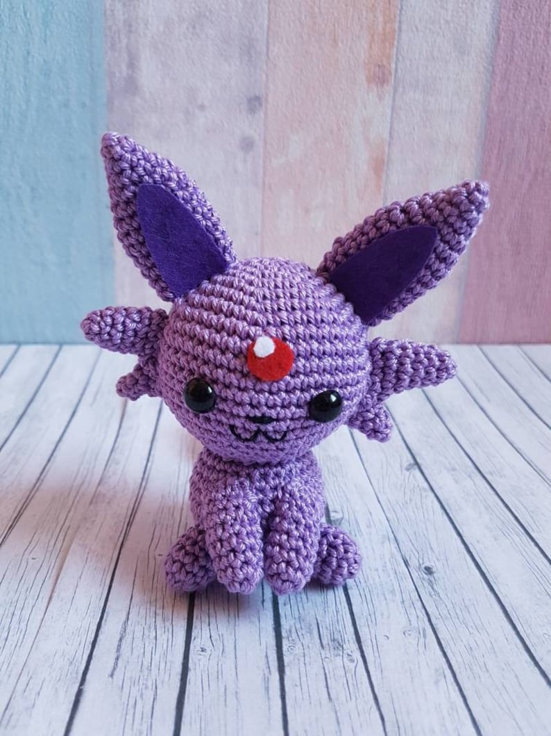 May include: Purple crocheted plush toy resembling a Pokemon character with large ears, a red dot on its forehead, and black eyes.