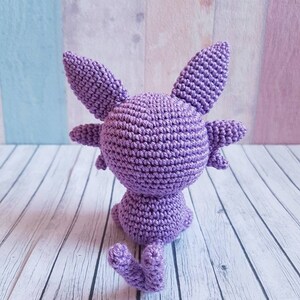 May include: A purple crocheted amigurumi toy in the shape of a cat sitting on a white wooden surface.